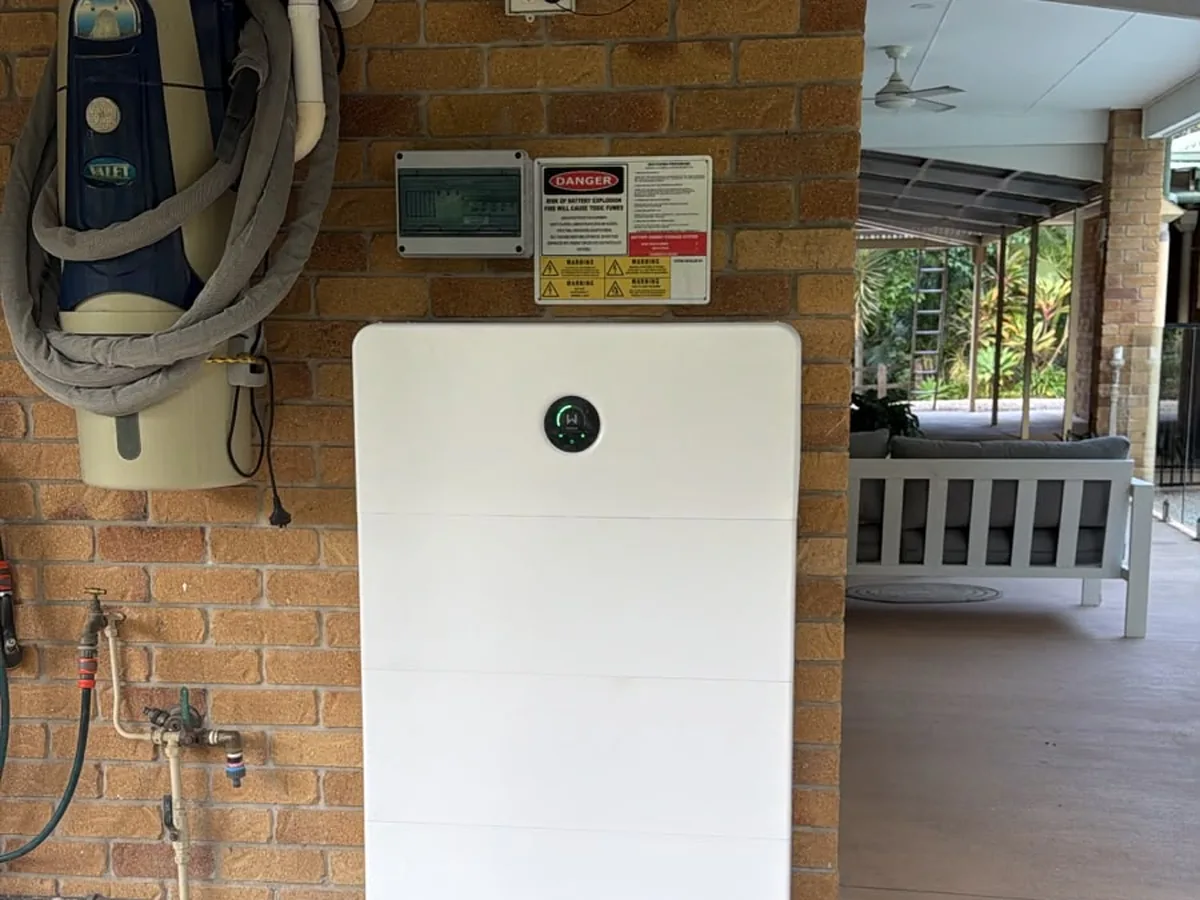 A home battery installed by EFS Solar against a brick wall under a Bonogin patio, beside the switchboard and shutdown signage