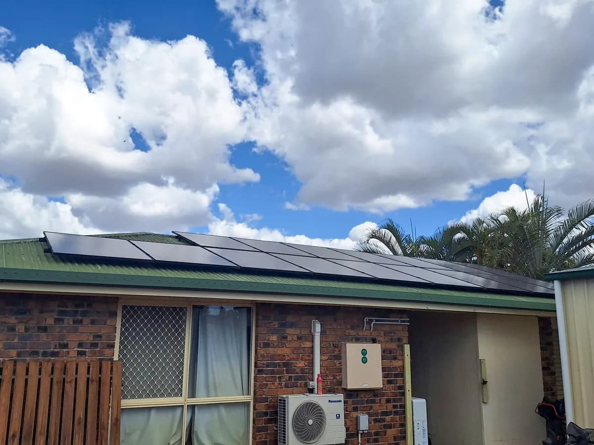 Black solar panels along the green metal roof of a brick home in Bundaberg East, with palms behind