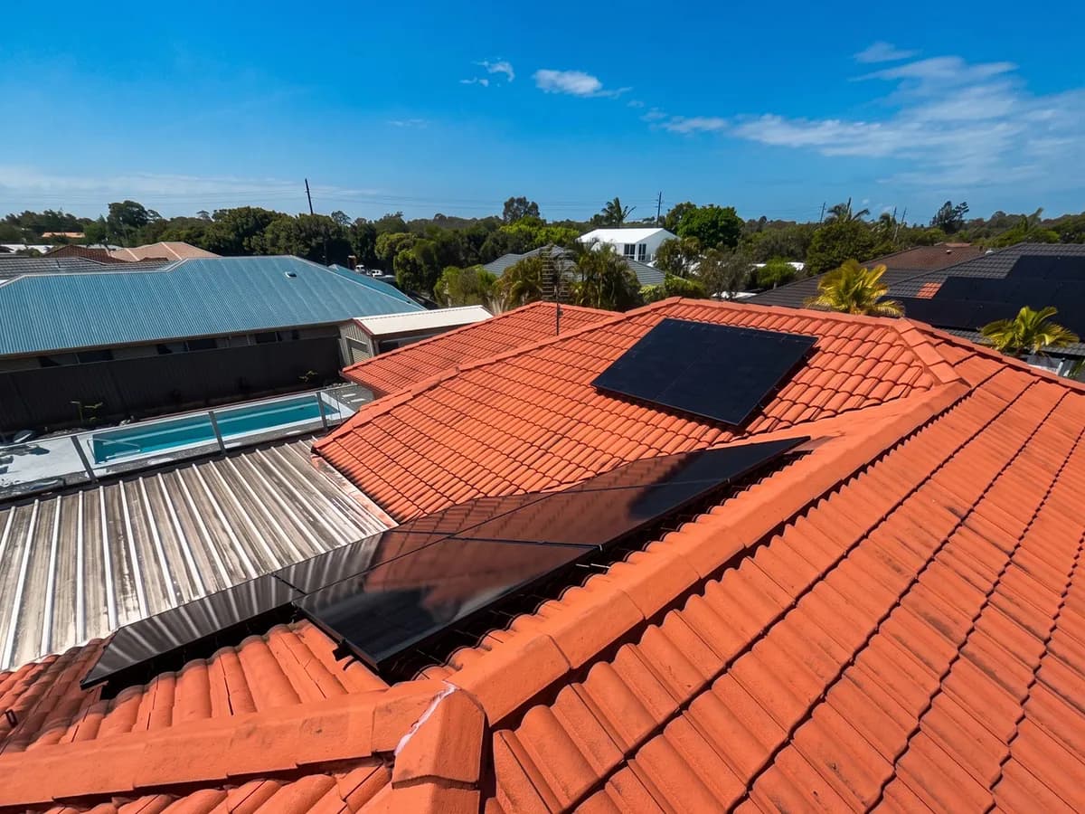 Aerial view of black solar panels on the red-tiled roof of a Currimundi home, with a pool and the neighbourhood behind