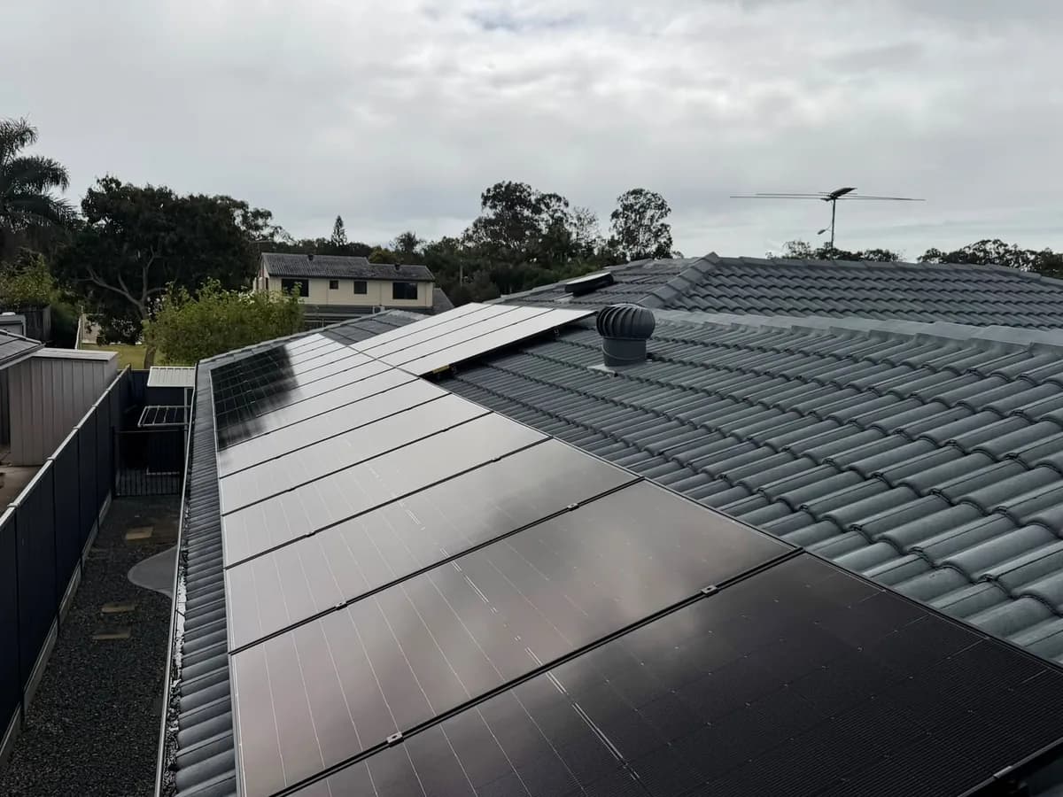 Black solar panels running along a grey tiled roof in Deception Bay, with a roof vent and the estate behind