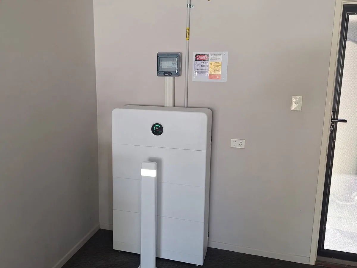 A home battery installed by EFS Solar indoors against a grey wall in Holmview, with the meter and shutdown signage above and a protective bollard in front