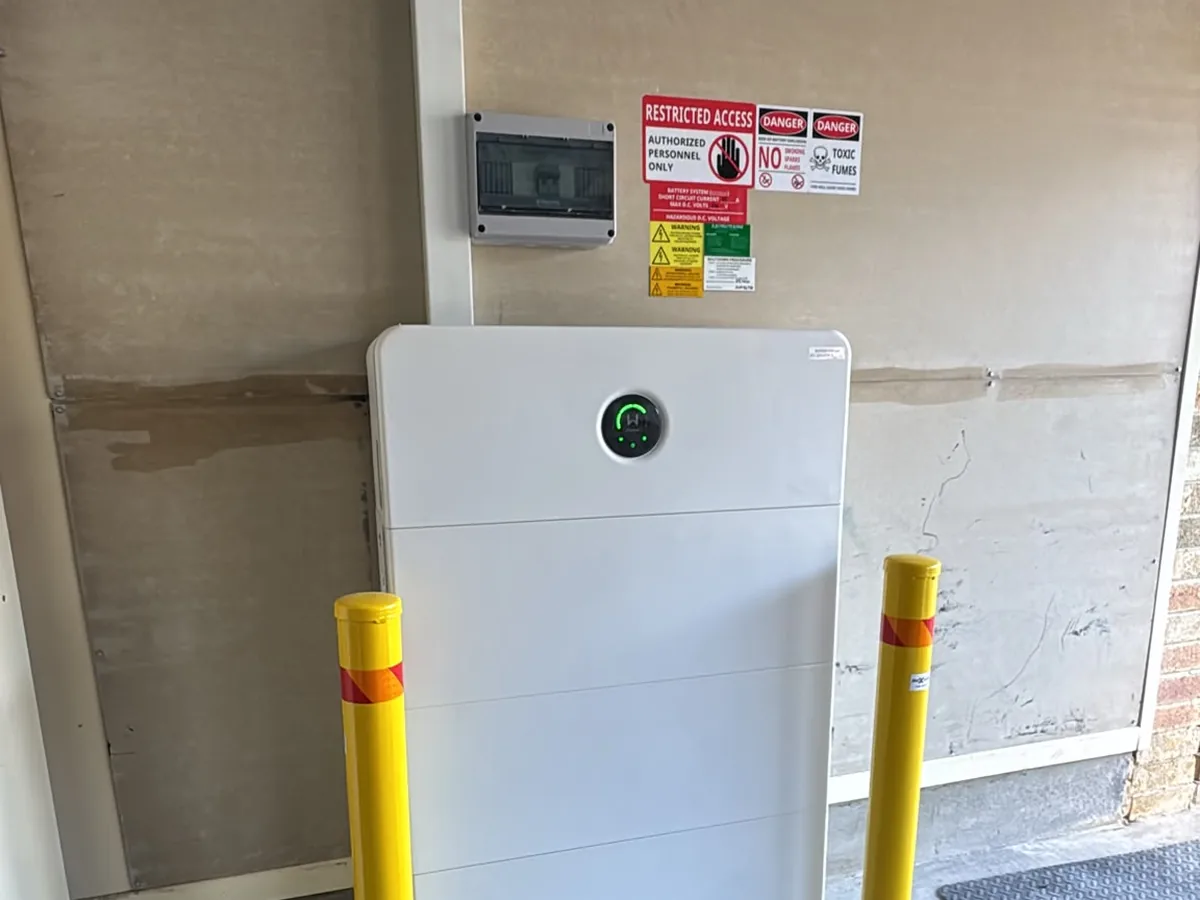 A home battery installed by EFS Solar in a Horsley garage, flanked by safety bollards, with the restricted-access and shutdown signage above