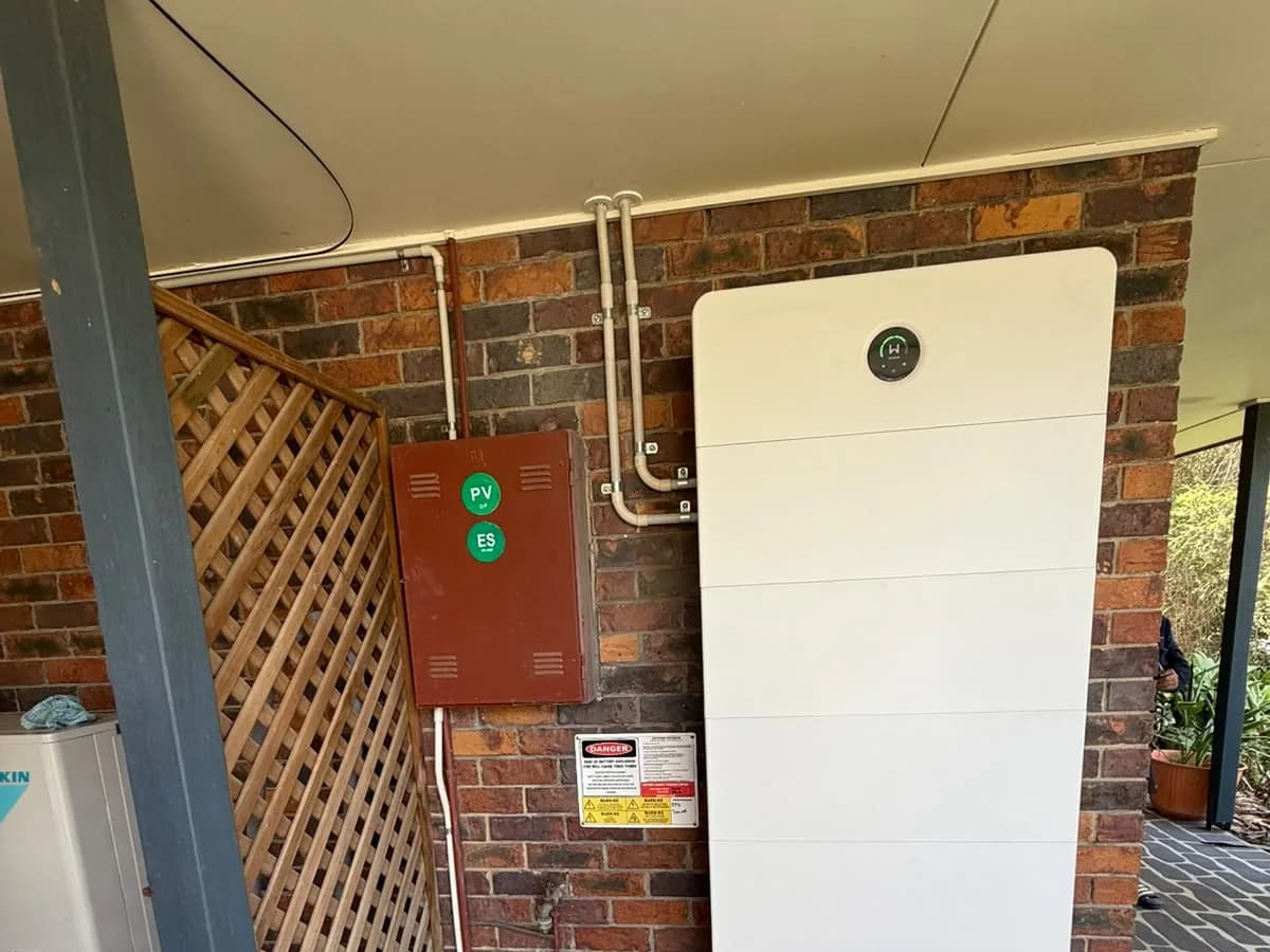 A tall home battery installed by EFS Solar against a brick wall in Karana Downs, beside the PV and battery isolators and shutdown signage