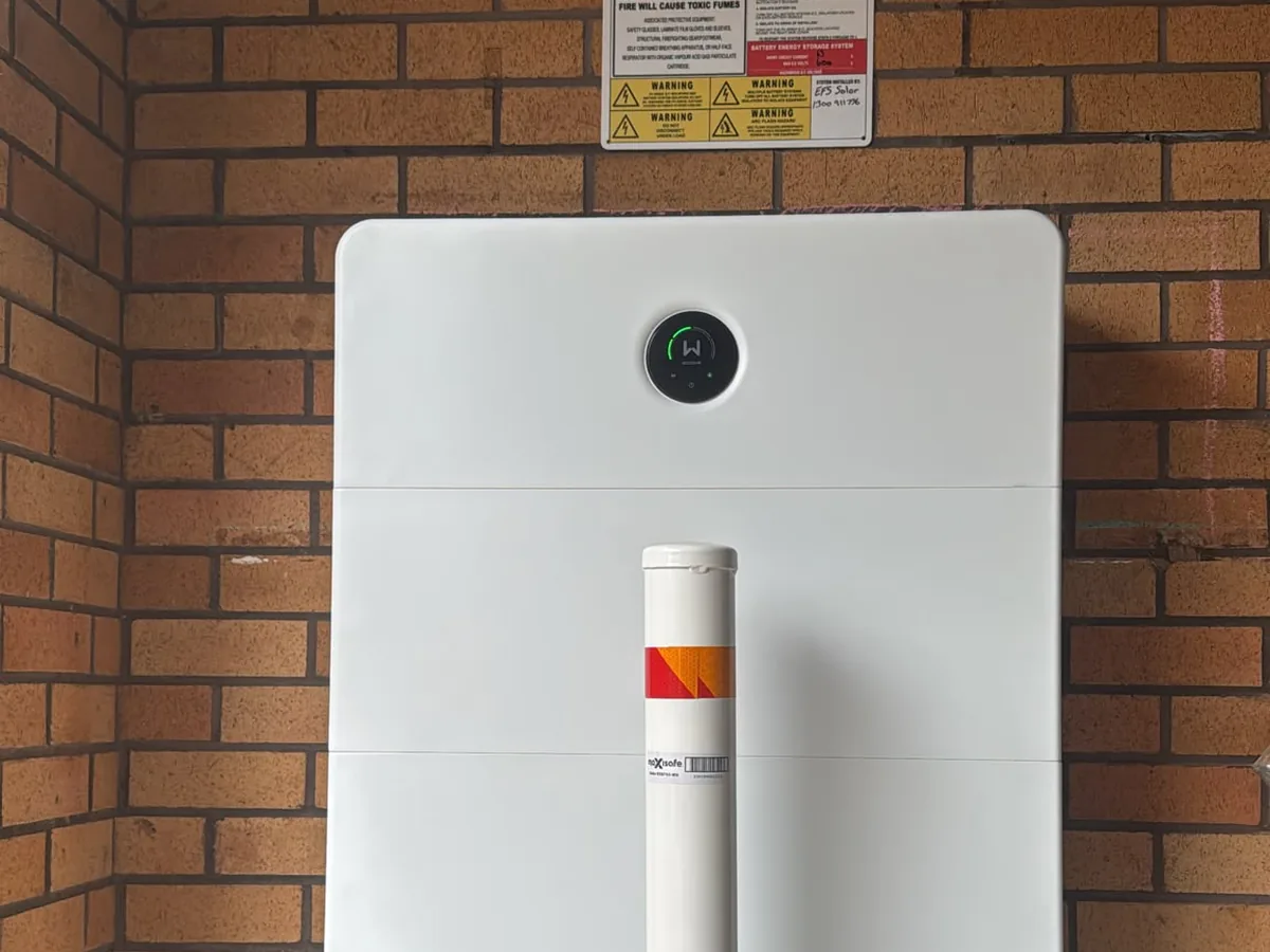 A home battery installed by EFS Solar against a brick wall in Macksville, with a protective bollard and shutdown signage