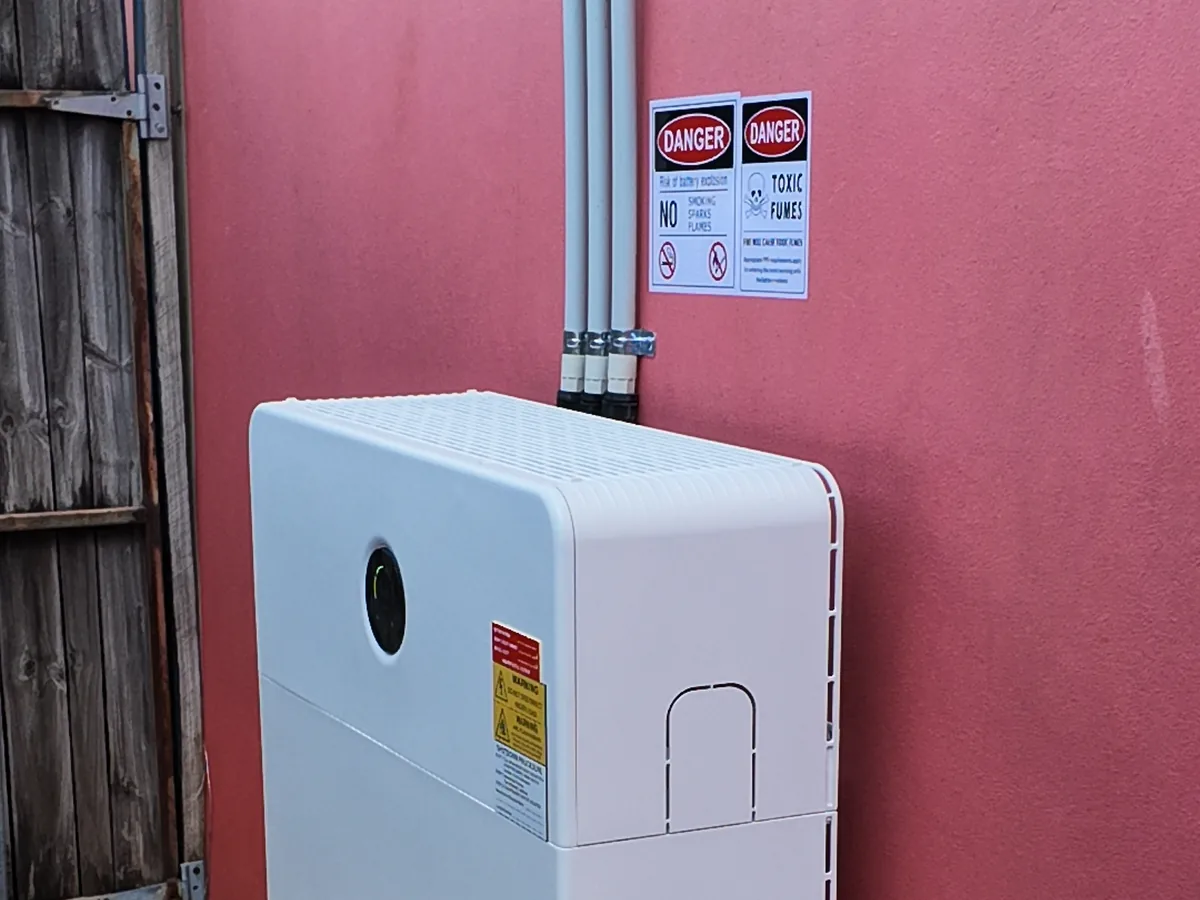 The top of a home battery installed by EFS Solar against a red rendered wall in Molendinar, with the conduits and shutdown signage above
