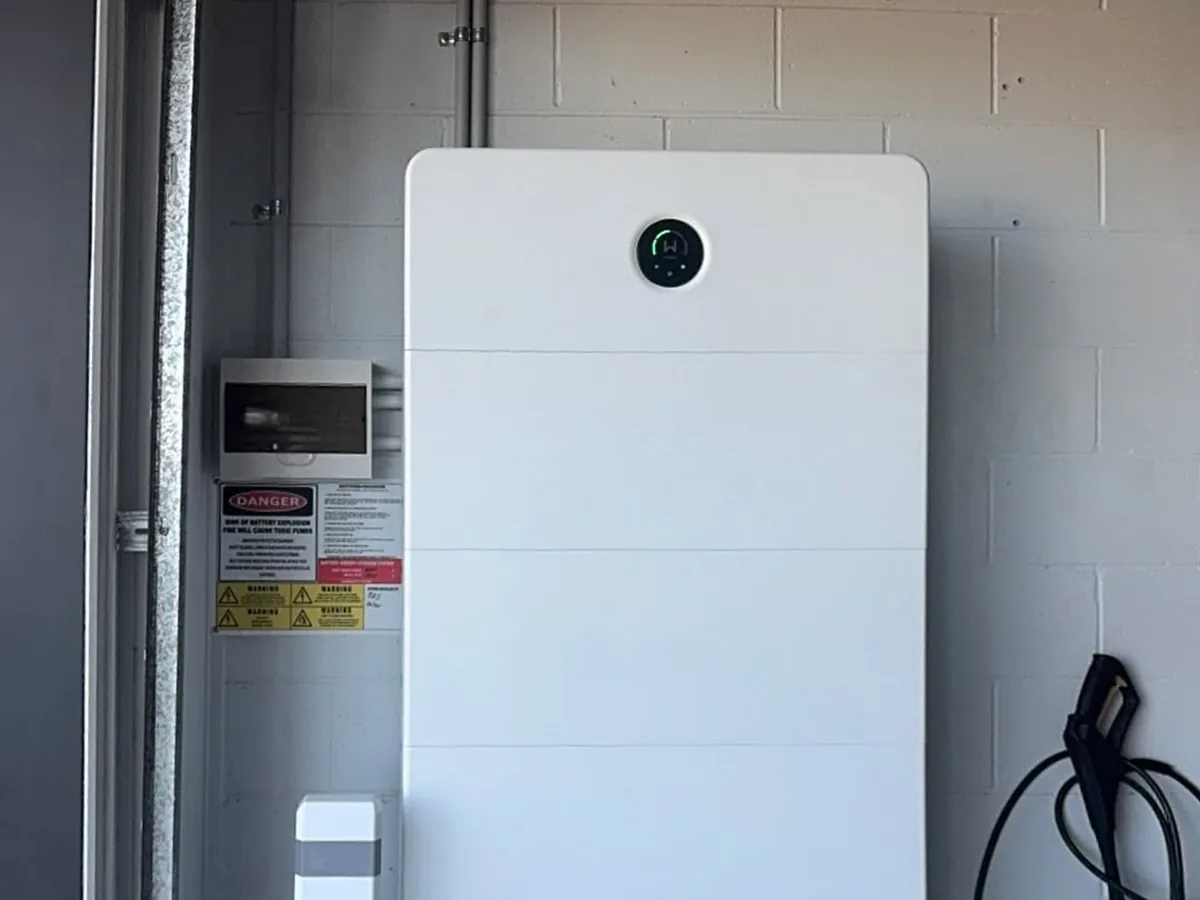 The top of a tall home battery installed by EFS Solar in a Morningside garage, with the switchboard and shutdown signage beside it