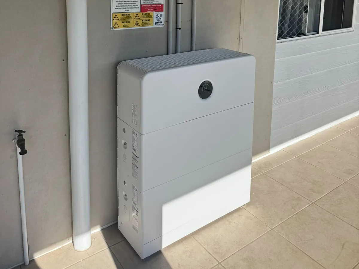 A home battery installed by EFS Solar against a rendered wall on a North Shore patio, beside the isolators and shutdown signage